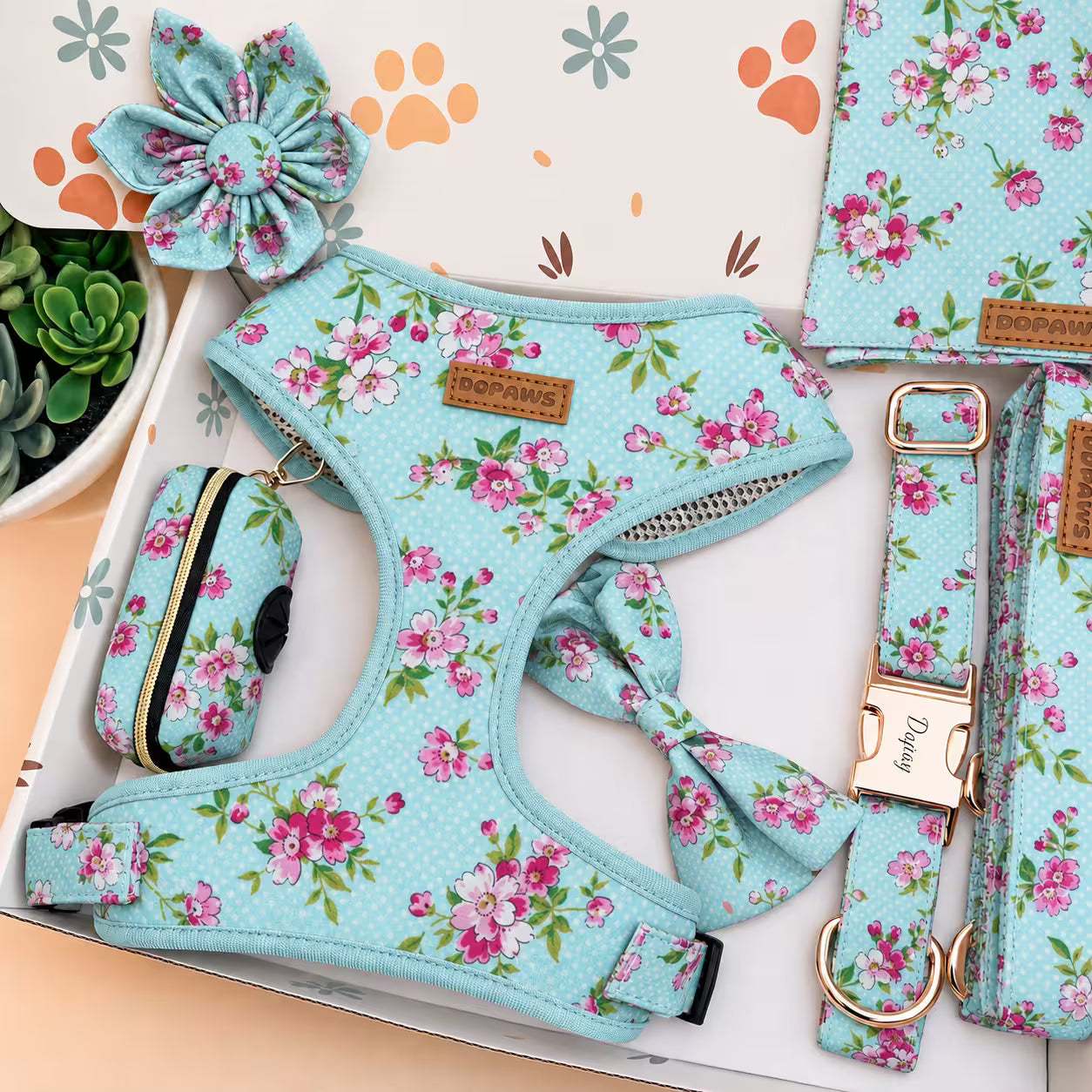 A pet accessory set in a floral print, featuring a light blue fabric harness, matching leash, small zippered pouch, and flower-shaped collar charm, all laid out in a box with a paw print patterned insert.