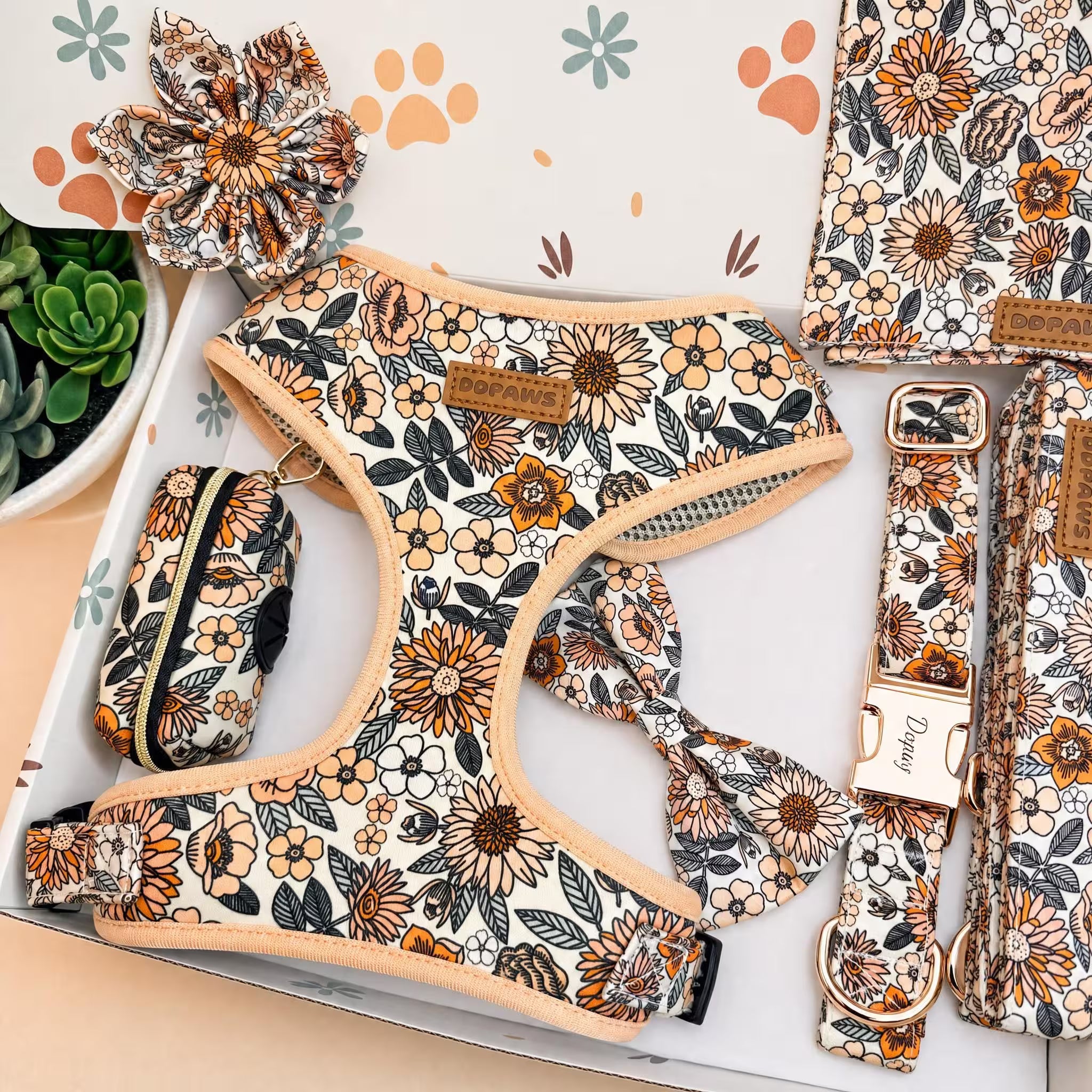 A pet accessory set laid out in a box, featuring a floral-print harness, matching leash, zip pouch, collar, and scrunchie. The set has a cream background with orange, brown, and black floral patterns, tan leather accents, and rose gold hardware.