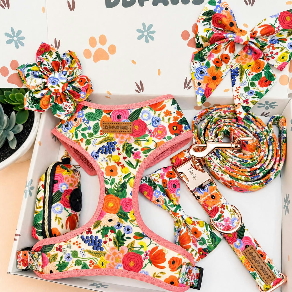 A colorful floral pet accessory set arranged in a white gift box, including a harness, leash, matching bow, collar, and small pouch. The fabric has a busy multicolor floral print with pink trim and a visible 'BOPAWS' brand patch.
