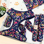 A dog gear set laid out on a white box, featuring a navy blue harness, matching collar, leash, and bow accessory, all printed with a colorful floral and bird pattern. A small leather brand patch is visible on the harness.