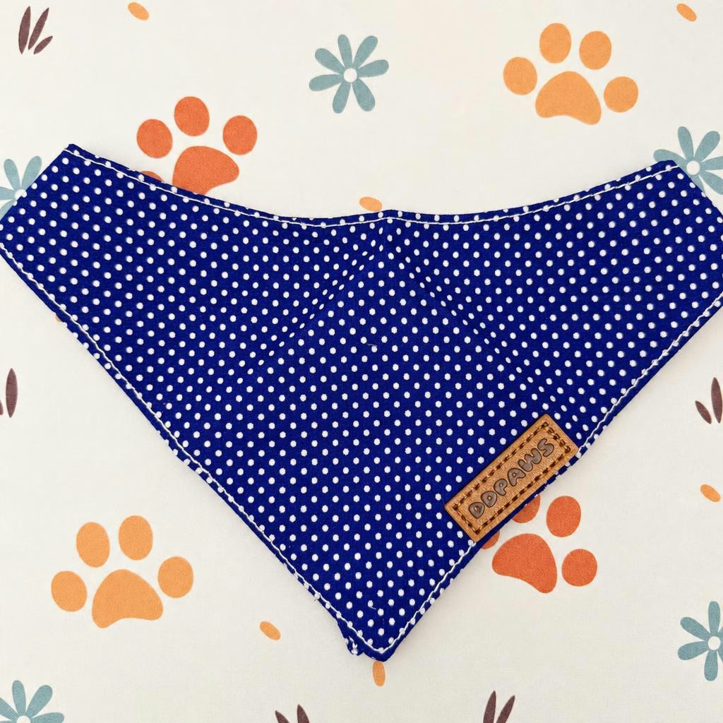 A blue and white polka dot pet bandana or collar laid flat on a white background with orange paw prints and blue flowers. A small tan fabric brand patch reading 'BERRYS' is sewn onto one corner.