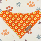 An orange dog bandana with a yellow, pink, and white floral pattern, featuring a brown fabric brand tag that reads 'COPOCALES'. The bandana is laid flat on a cream-colored background decorated with paw prints and flowers.