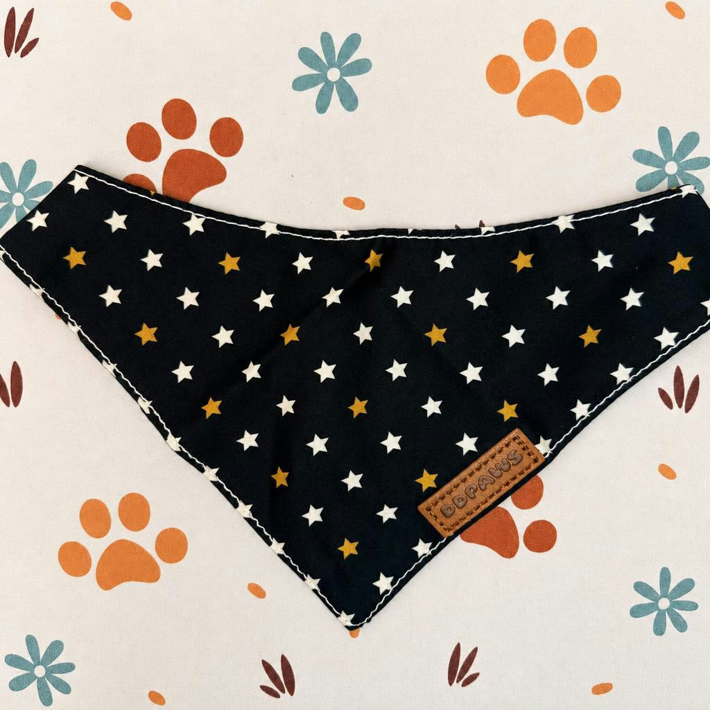A triangular dog bandana with a black background, decorated with white and gold stars, laid flat on a cream-colored background patterned with orange paw prints and blue flowers. A small brown leather brand tag is stitched onto the bottom right corner.