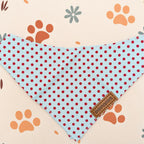 A light blue triangular pet bandana with red polka dots, featuring a small brown leather brand tag that reads 'LOVE PUPPUS'. The bandana is laid flat on a cream-colored background decorated with orange paw prints and small gray flowers.