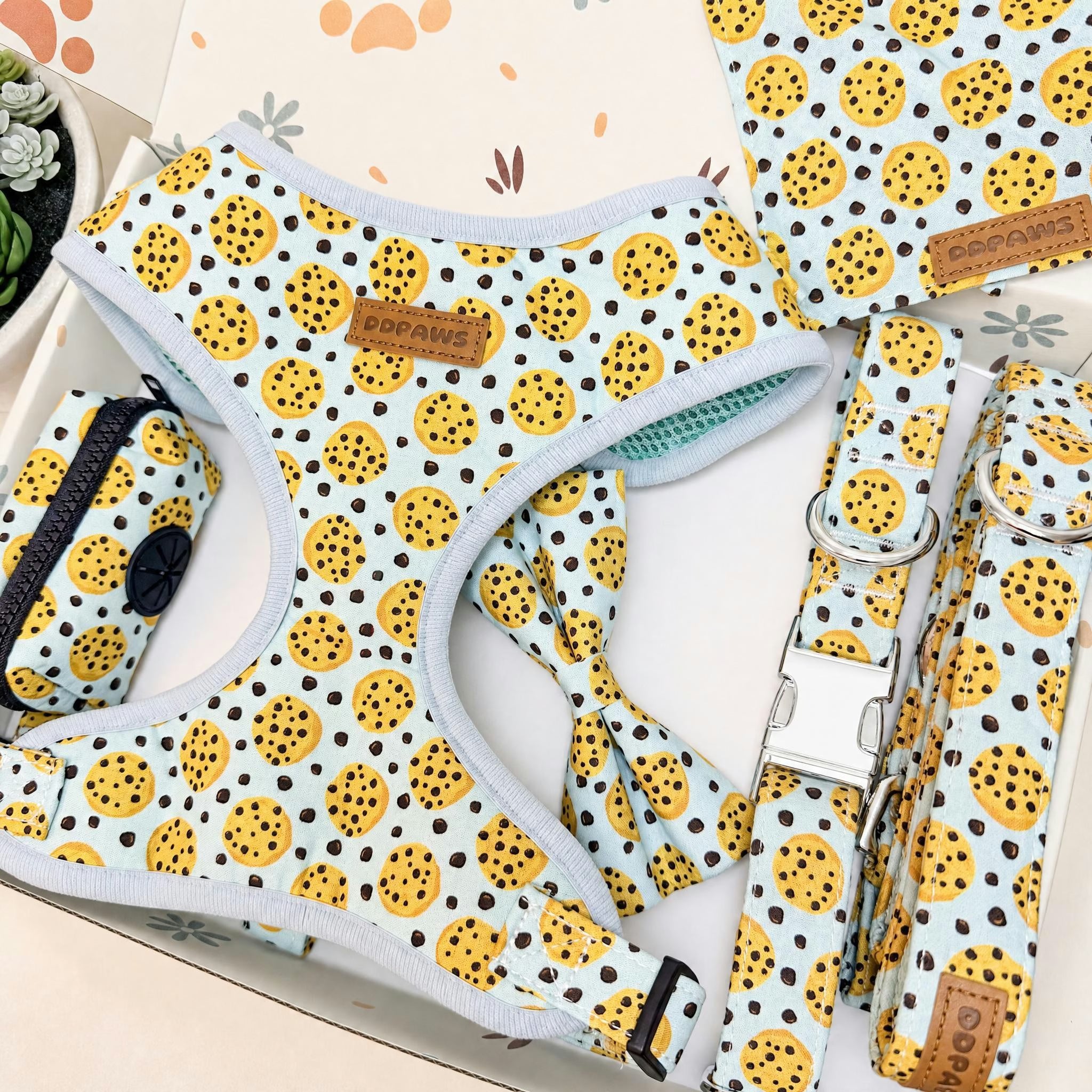 A dog harness set laid out on a light surface, featuring a matching harness, leash, and small pouch. The fabric has a light blue base with a yellow and black spotted pattern, and a small brown leather brand patch reading 'Pup Patterning'.