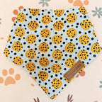 A triangular pet bandana bib with a light blue background, covered in a pattern of yellow cookies with black spots, laid flat on a cream-colored surface with paw prints and floral motifs. A brown leather brand tag reading 'Doggolux' is sewn onto the lower right corner.