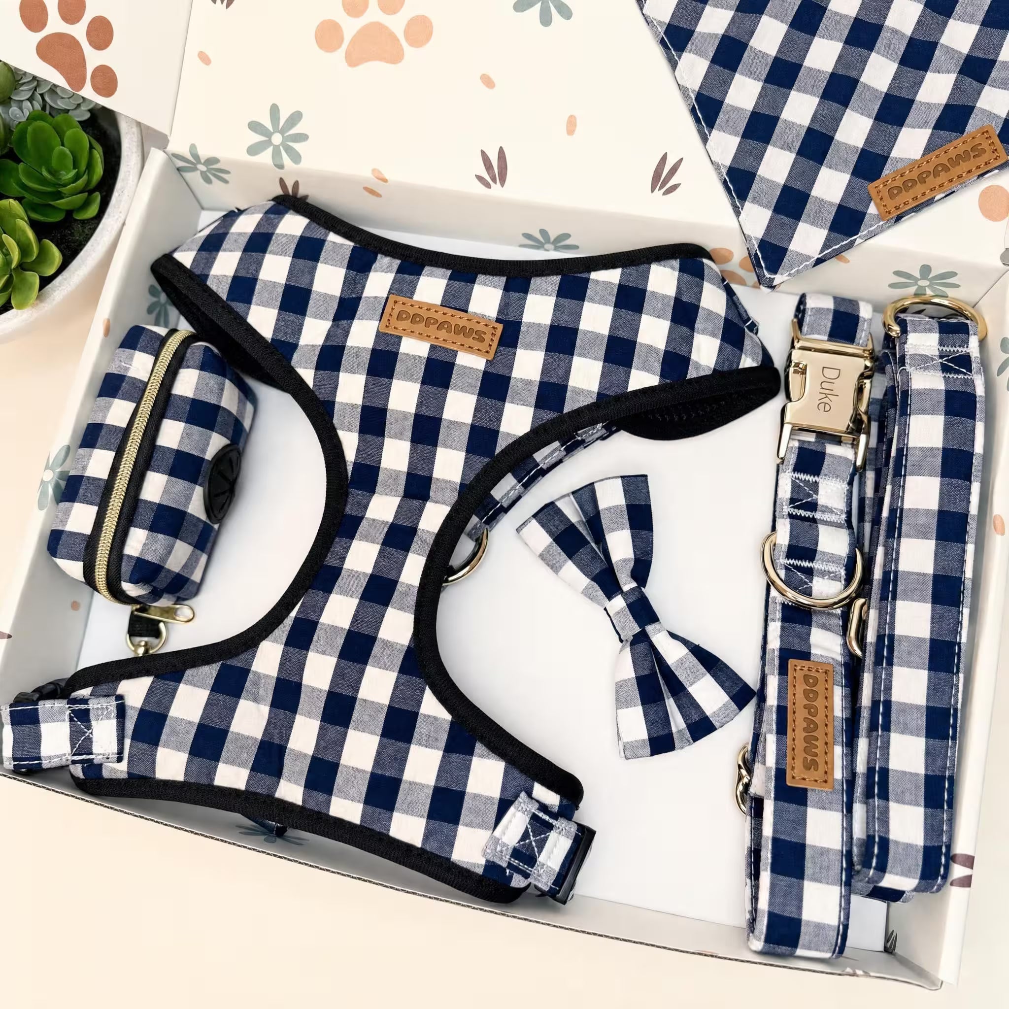 A blue and white gingham check pet accessory set displayed inside a white gift box, including a harness, leash, collar, and matching bow tie, with a small plant in the corner.